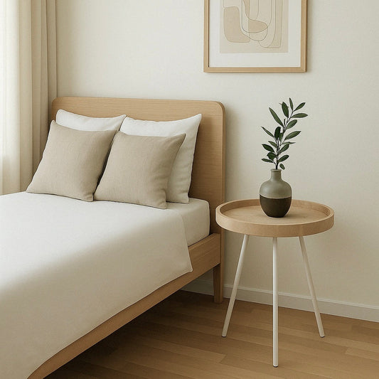 Skandinavisches Schlafzimmer mit einem Bett aus hellem Holz, weißen und beigen Kissen sowie einem runden Nachttisch mit schlanken, weißen Metallbeinen. Auf dem Tisch steht eine Keramikvase mit einem Olivenzweig, natürlich beleuchtet durch Tageslicht